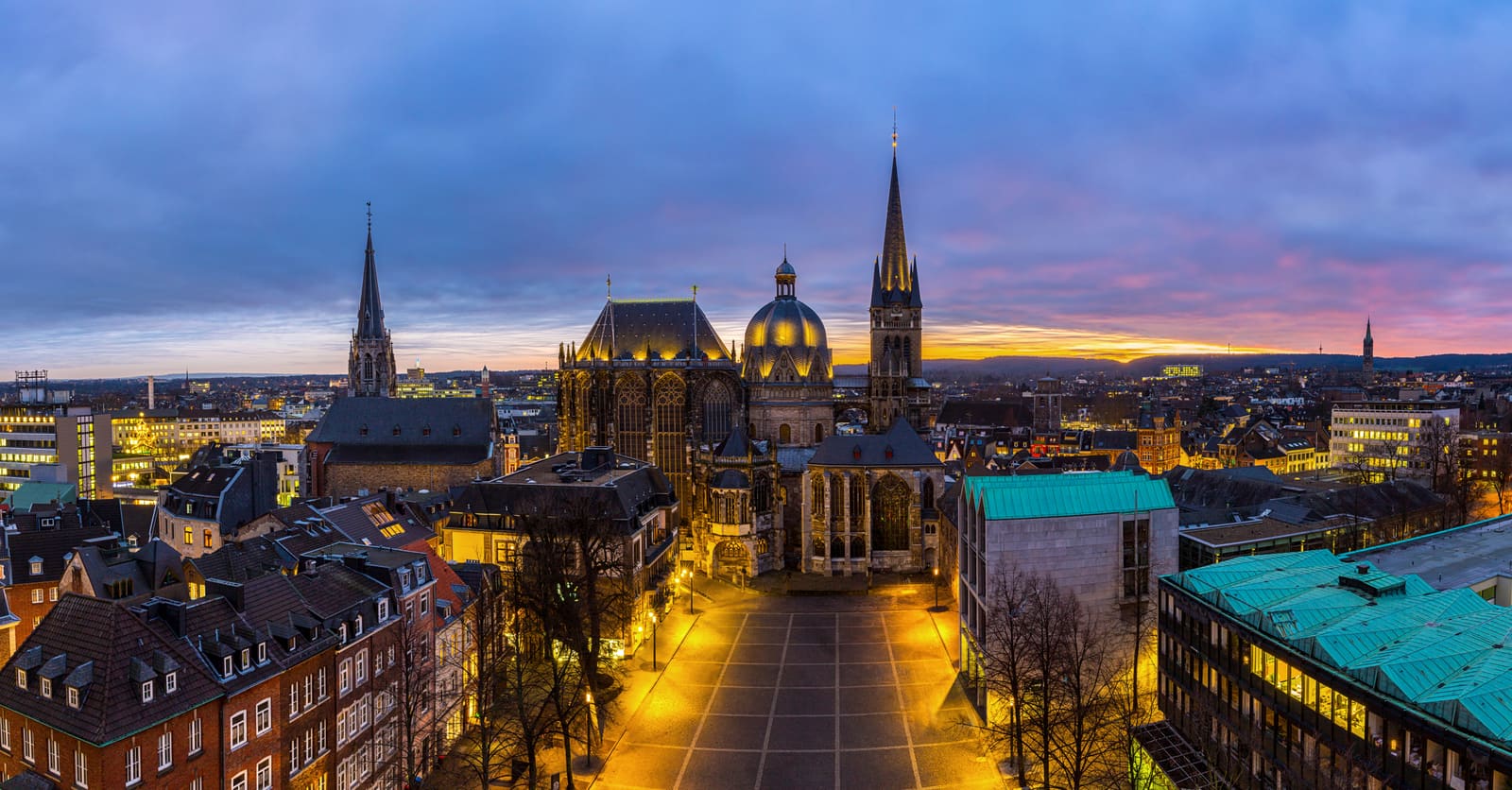 Beleuchteter Aachener Dom mit Oktogon, Chorhalle und Westturm über den Dächern der Altstadt bei farbigem Sonnenuntergang
