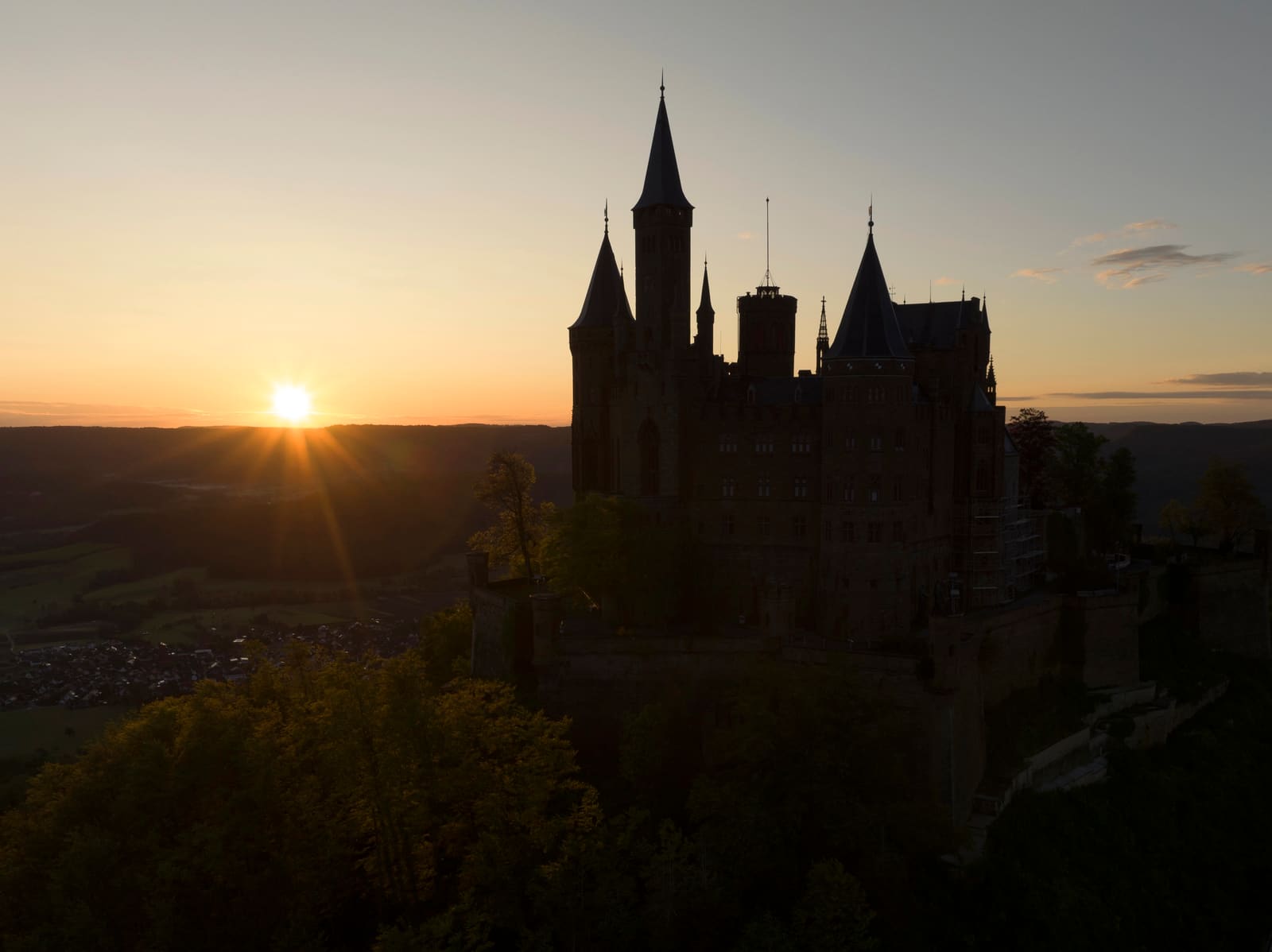 Burg Hohenzollern als dunkle Silhouette auf dem bewaldeten Bergkegel neben der tief stehenden Morgensonne