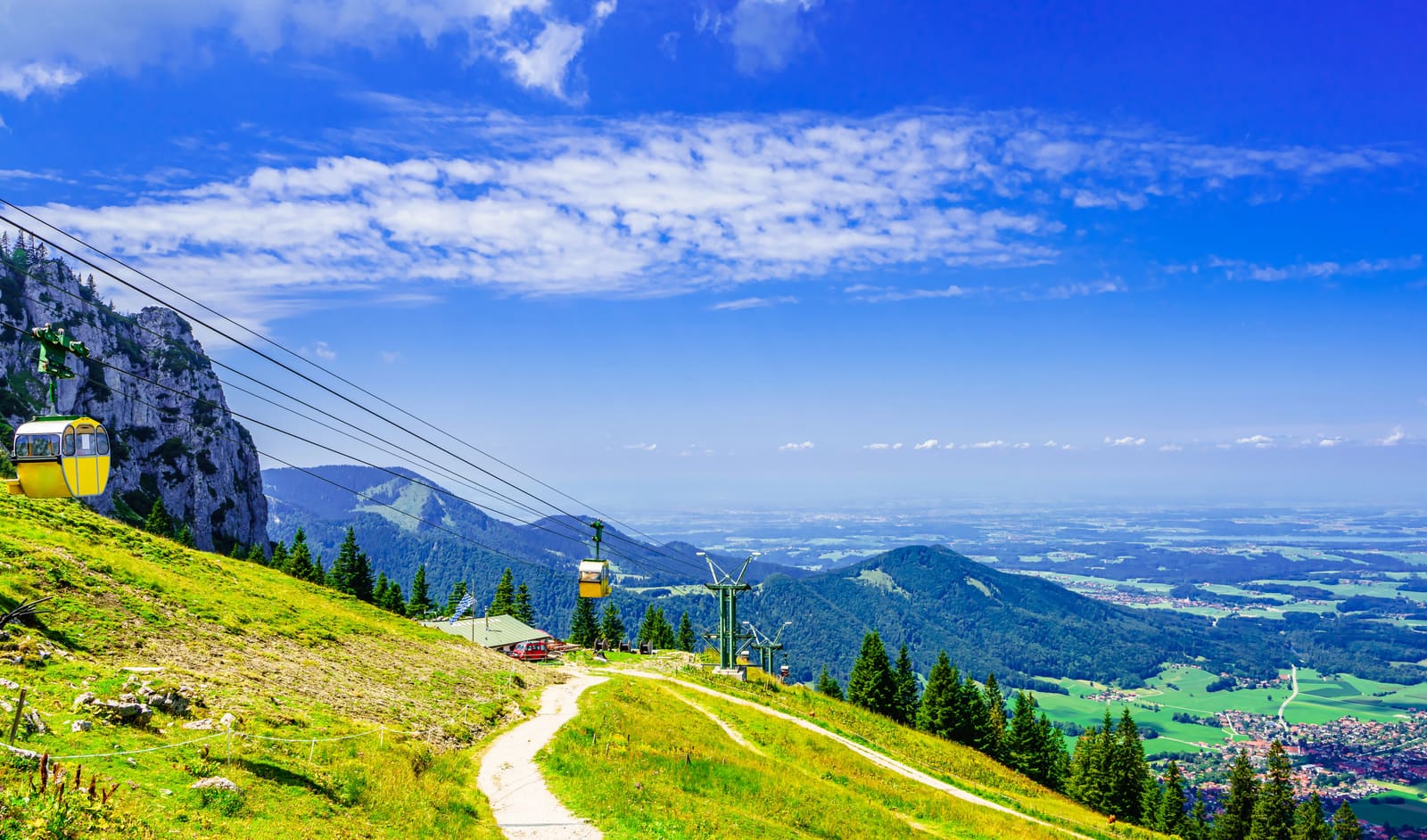 Gelbe Gondeln der Kampenwandbahn über einem Wanderweg, dahinter der weite Blick ins Chiemgauer Voralpenland