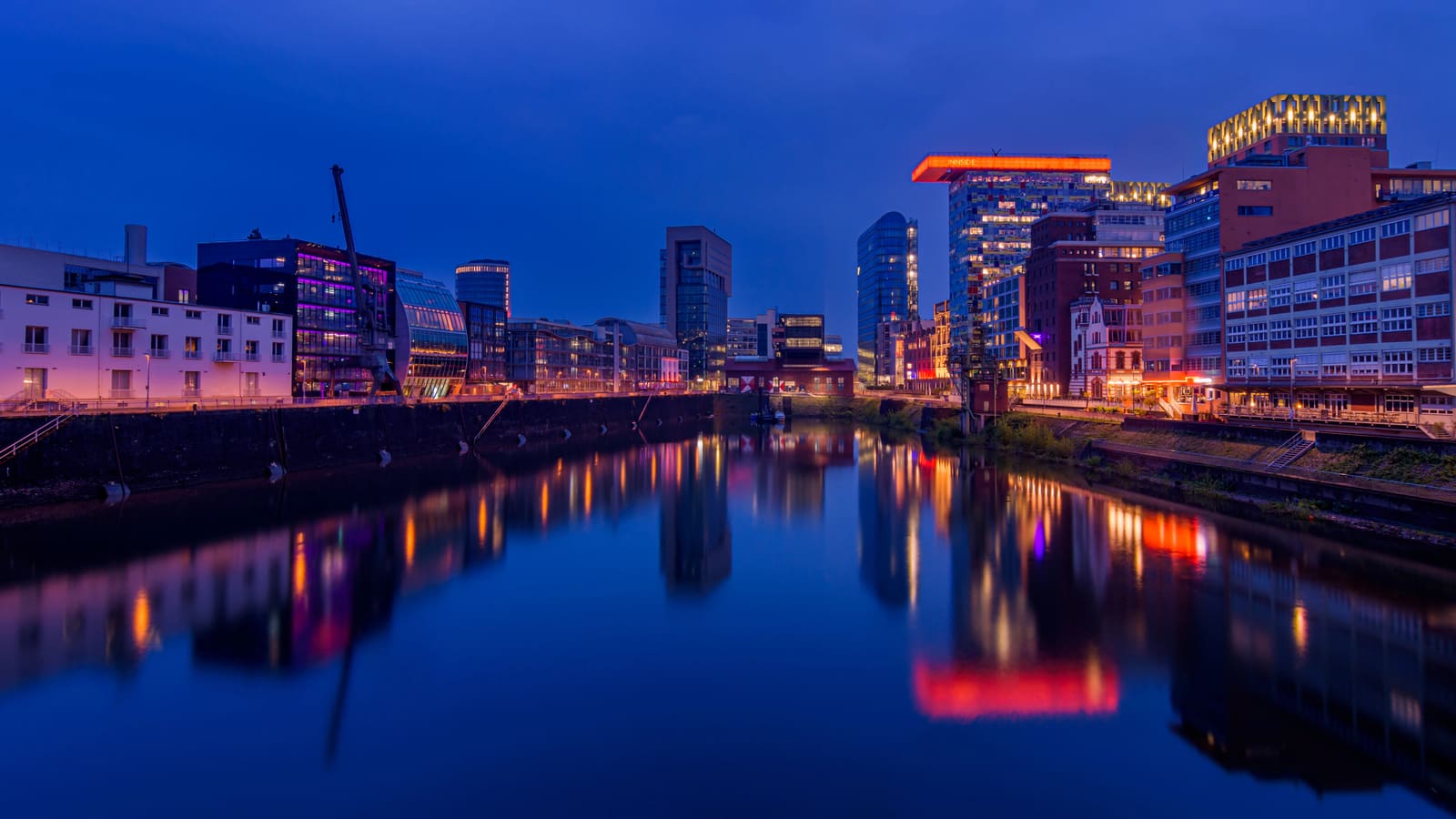 Beleuchtete moderne und historische Gebäude im Düsseldorfer Medienhafen mit farbigen Spiegelungen im ruhigen Hafenbecken