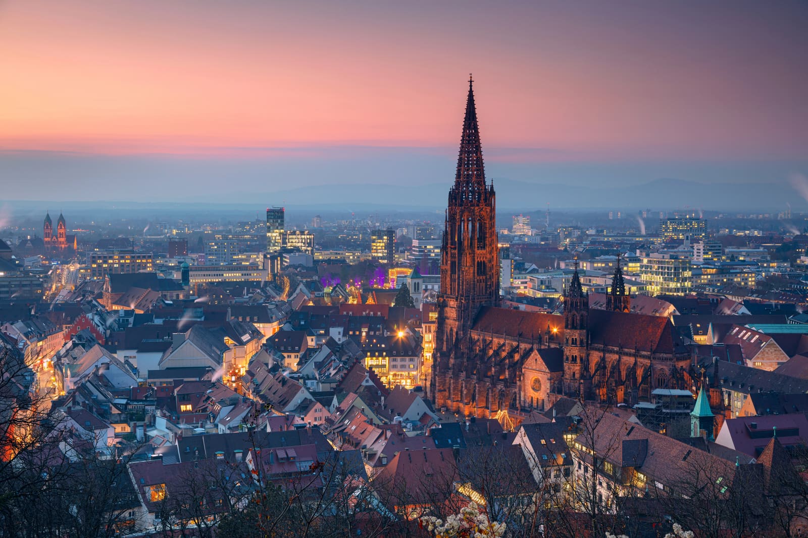 Freiburger Münster mit seinem durchbrochenen gotischen Turm über der beleuchteten Altstadt und den fernen Schwarzwaldhöhen am Abend