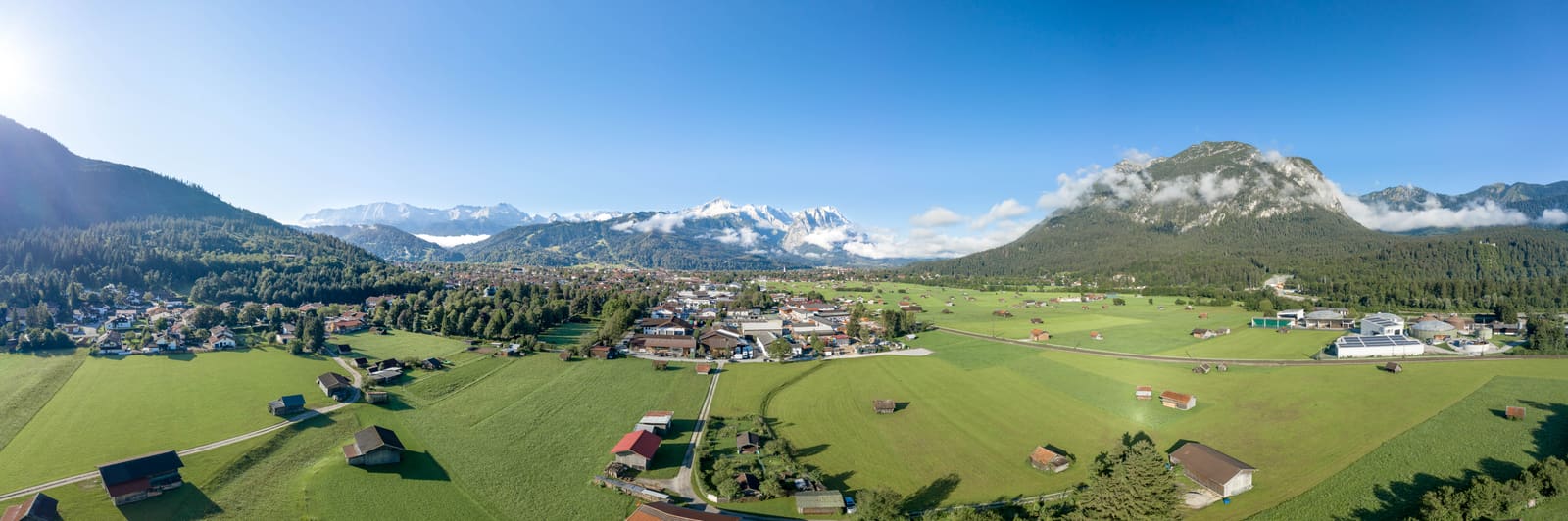 Luftpanorama von Garmisch-Partenkirchen mit grünen Wiesen, verstreuten Heustadeln und bewaldeten Bergen unter blauem Himmel
