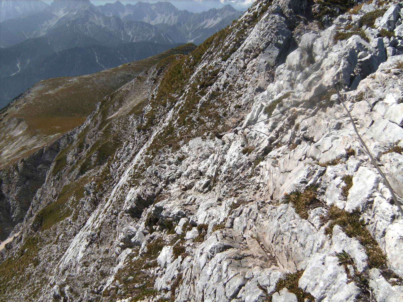 Schmaler Mittenwalder Klettersteig mit Stahlseil an einer steilen hellen Felswand und tiefem Blick ins Karwendel