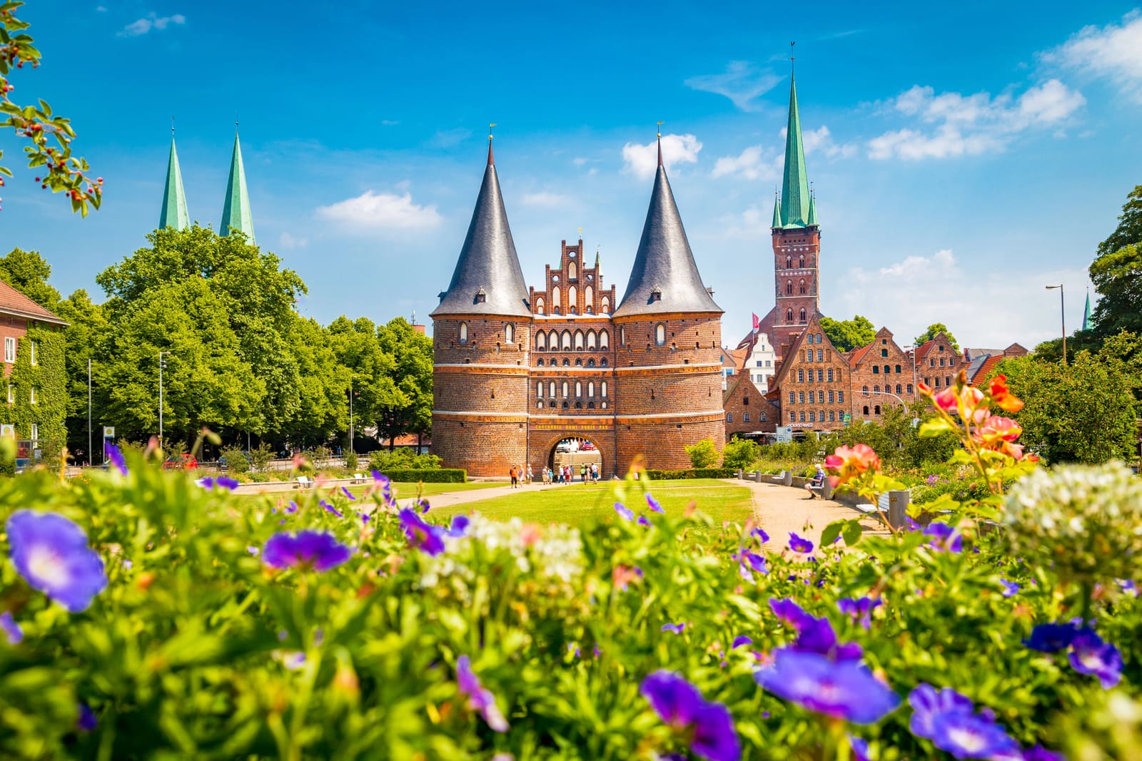 Lübecker Holstentor mit seinen beiden Rundtürmen vor Altstadtgiebeln und Kirchtürmen, eingerahmt von sommerlichen Blumen