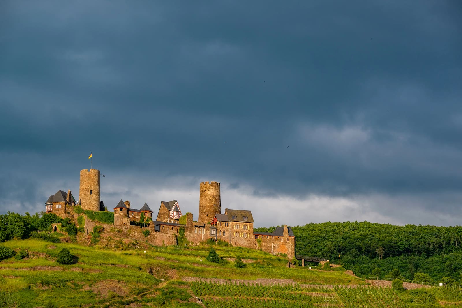 Burg Thurant mit ihren beiden Rundtürmen über grünen Weinbergen bei Alken unter einem dramatisch dunklen Himmel