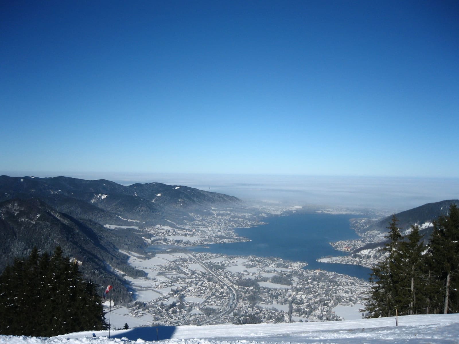 Blick vom verschneiten Wallberg über den Tegernsee unter klarem Winterhimmel.